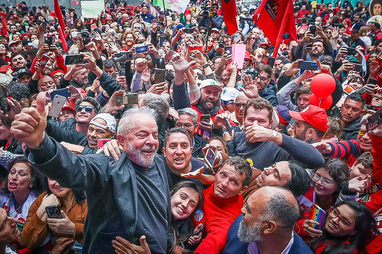 Lula sorri e faz sinal de positivo, cercado por apoiadores com celulares, bandeiras vermelhas e balões em ato de campanha
